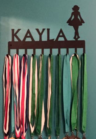 A medal hanger with a metal silhouette of an Irish dancer and the name KAYLA. It has 10 hooks with medals hanging from them. It is made in the USA.