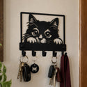 A black metal wall-mounted key rack with a cat face silhouette at the top and several hanging keys and a burgundy strap, shown on a beige wall in a real interior setting.