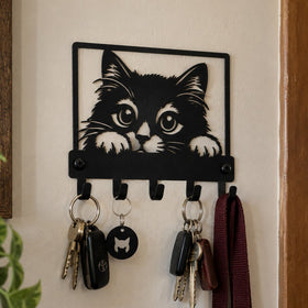 A black metal wall-mounted key rack with a cat face silhouette at the top and several hanging keys and a burgundy strap, shown on a beige wall in a real interior setting.