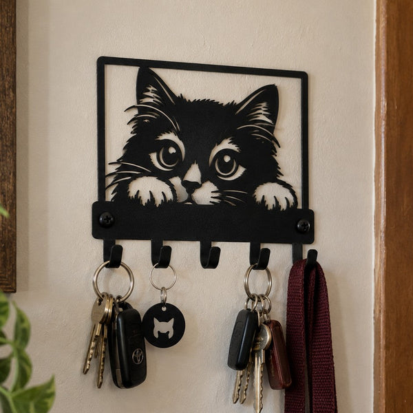 A black metal wall-mounted key rack with a cat face silhouette at the top and several hanging keys and a burgundy strap, shown on a beige wall in a real interior setting.