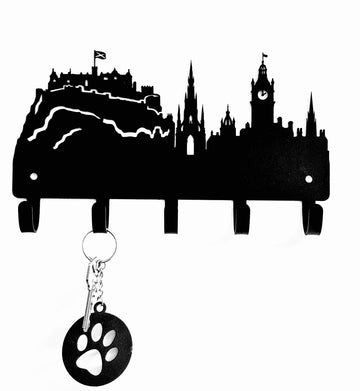 Black metal wall key holder shaped like a city skyline with five hanging hooks, shown on a white background with a sample key and paw-shaped tag hanging from one hook.