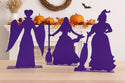 Silhouettes of 3 purple witches on a table with Halloween decorations in the background.