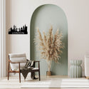 A black metal London skyline wall art piece with the word 'LONDON' is displayed on a beige wall in a styled living room setting featuring a green arched accent wall, a wooden armchair, and a pampas grass arrangement.