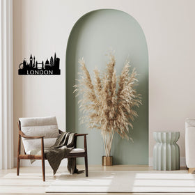 A black metal London skyline wall art piece with the word 'LONDON' is displayed on a beige wall in a styled living room setting featuring a green arched accent wall, a wooden armchair, and a pampas grass arrangement.