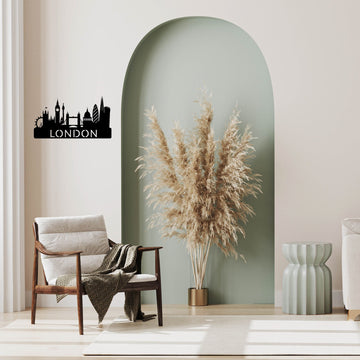 A black metal London skyline wall art piece with the word 'LONDON' is displayed on a beige wall in a styled living room setting featuring a green arched accent wall, a wooden armchair, and a pampas grass arrangement.