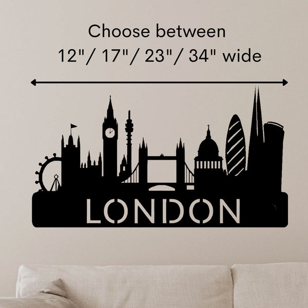 Black London skyline wall decal with silhouettes of famous landmarks and the word 'LONDON' at the bottom, shown on a beige wall above a sofa. Text above reads 'Choose between 12" / 17" / 23" / 34" wide' with a measurement arrow.