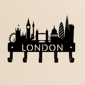 A black metal London skyline wall decor piece with five hooks at the bottom, featuring silhouettes of landmarks including Big Ben, Tower Bridge, and the London Eye on a beige background.