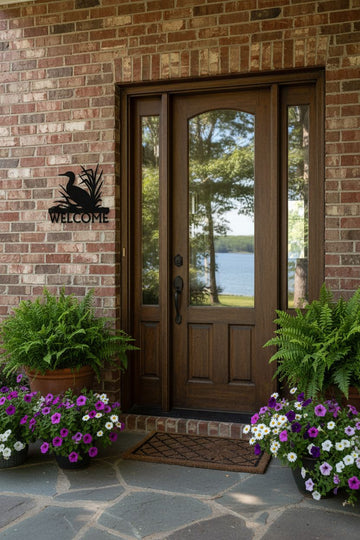 A front entryway with dark brown wooden double doors featuring glass panels, set in a brick wall with potted ferns, purple and white flowers, and a stone walkway. A black metal 'WELCOME' sign with a duck silhouette is mounted on the brick wall.