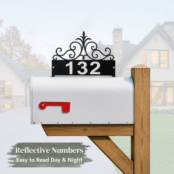 A white mailbox mounted on a wooden post with a decorative black metal sign above the door displaying the number 132 in reflective white numerals. The background shows a blurred suburban house and lawn.