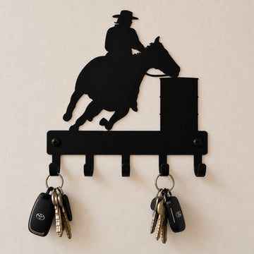 A black metal wall-mounted key rack shaped like a cowboy riding a horse over a barrel, shown on a light beige wall with two keys hanging from the hooks.