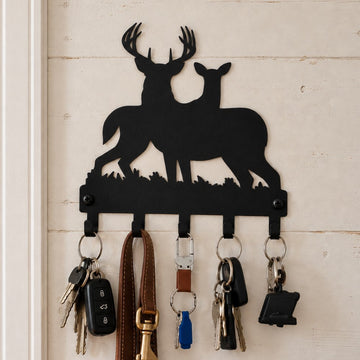 A black metal key holder shaped like two deer silhouettes hangs on a white wood-paneled wall. Several keys and a brown leather leash are attached to the five hooks below.