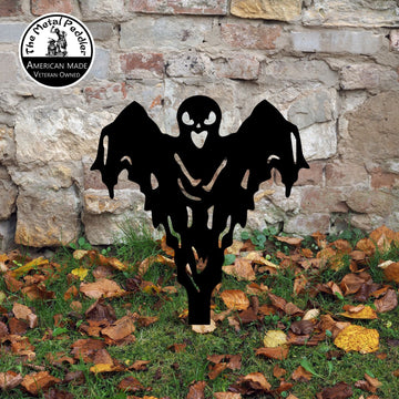 Black ghost-shaped decorative yard art in front of a stone wall with fallen leaves on the ground.