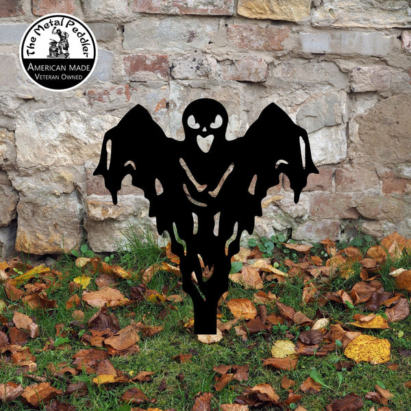 Black ghost-shaped decorative yard art in front of a stone wall with fallen leaves on the ground.