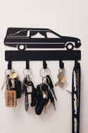 A black metal wall-mounted key holder shaped like a hearse with five hanging hooks holding various keys and a vertical organizer strip on the right, shown against a plain white wall.
