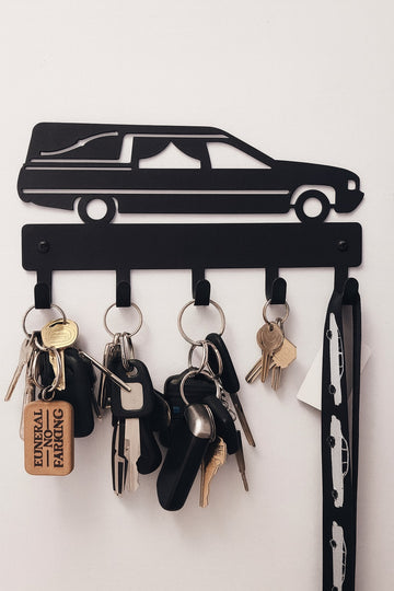 A black metal wall-mounted key holder shaped like a hearse with five hanging hooks holding various keys and a vertical organizer strip on the right, shown against a plain white wall.