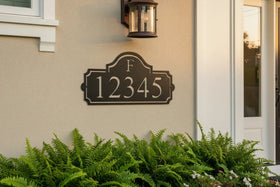 A dark metal house number plaque with the letters 'F' and numbers '12345' in raised silver lettering, mounted on a beige stucco wall above green ferns next to a white door.
