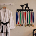 A white martial arts uniform with a black belt hanging on a wooden hanger next to a black wall-mounted display holding multiple medals with colorful ribbons.