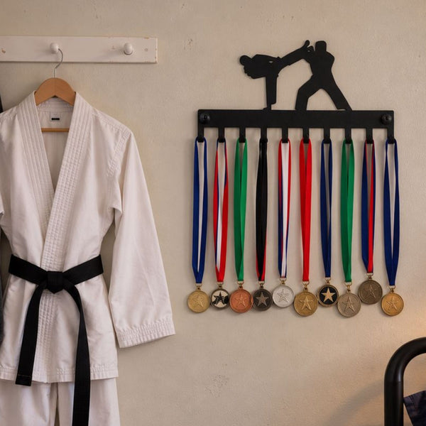A white martial arts uniform with a black belt hanging on a wooden hanger next to a black wall-mounted display holding multiple medals with colorful ribbons.