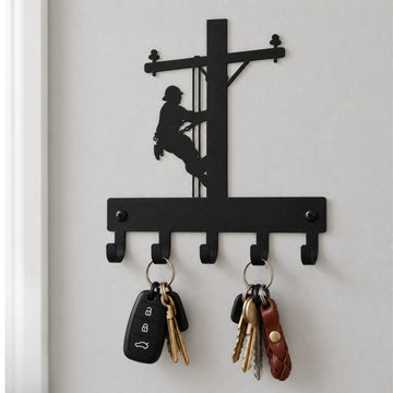 Black metal wall hook rack shaped like a utility pole with a lineman silhouette climbing the pole, mounted on a light gray wall. Two keys hang from the hooks, and a white door frame is visible on the left.
