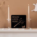A black sign that says Magic Lives Here sitting on a mantelpiece next to 2 candlesticks