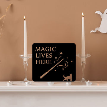 A black sign that says Magic Lives Here sitting on a mantelpiece next to 2 candlesticks