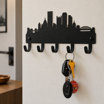 A black metal wall hook rack shaped like a city skyline, mounted on a white wall. Five hooks are visible, with a set of keys hanging from the center hook.
