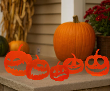 Set of orange pumpkin-shaped cutouts with carved faces on a stone surface, with pumpkins and flowers in the background.