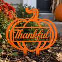 An orange metal yard sign shaped like a pumpkin with the word 'Thankful' in script across the center, displayed outdoors in a garden bed with flowers, soil, and a pumpkin nearby.