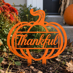An orange metal yard sign shaped like a pumpkin with the word 'Thankful' in script across the center, displayed outdoors in a garden bed with flowers, soil, and a pumpkin nearby.