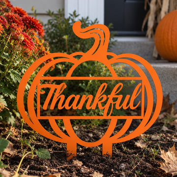 An orange metal yard sign shaped like a pumpkin with the word 'Thankful' in script across the center, displayed outdoors in a garden bed with flowers, soil, and a pumpkin nearby.