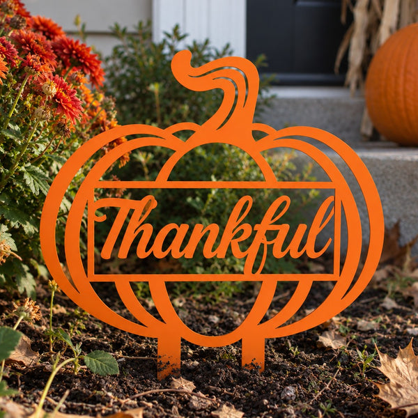 An orange metal yard sign shaped like a pumpkin with the word 'Thankful' in script across the center, displayed outdoors in a garden bed with flowers, soil, and a pumpkin nearby.