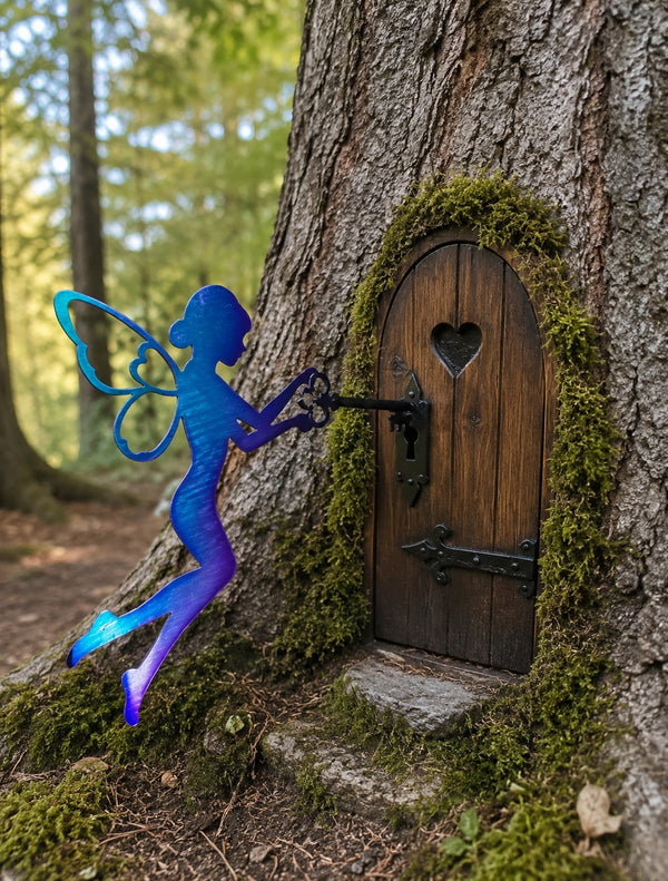 A blue and purple fairy silhouette garden stake stands beside a mossy tree trunk with a small wooden fairy door and key in a forest setting.